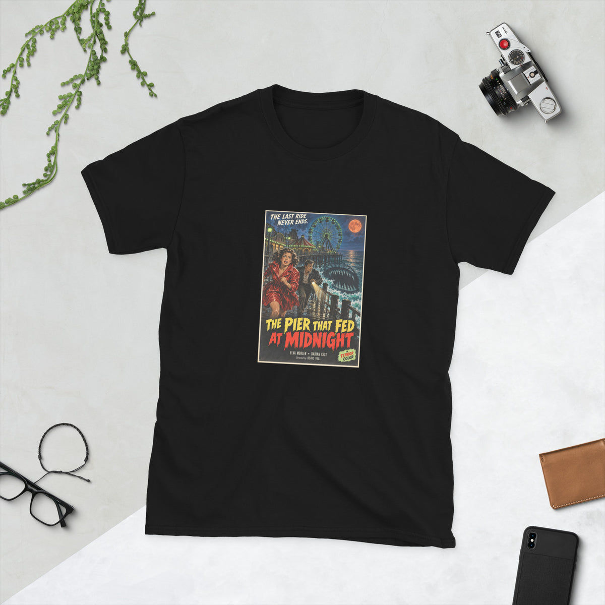 Black t-shirt printed with a vintage-style horror poster: a woman in a red raincoat and a man with a flashlight running along a pier at night past a lit Ferris wheel, a toothed shadow in the water, titled The Pier That Fed at Midnight