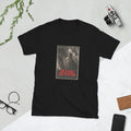 Black t-shirt printed with a sepia horror movie poster of a cracked plaster mask hanging by twine from a staircase post, with two more masks and a jack-o'-lantern in the shadows below, under the red title "The Faces Are Waiting."