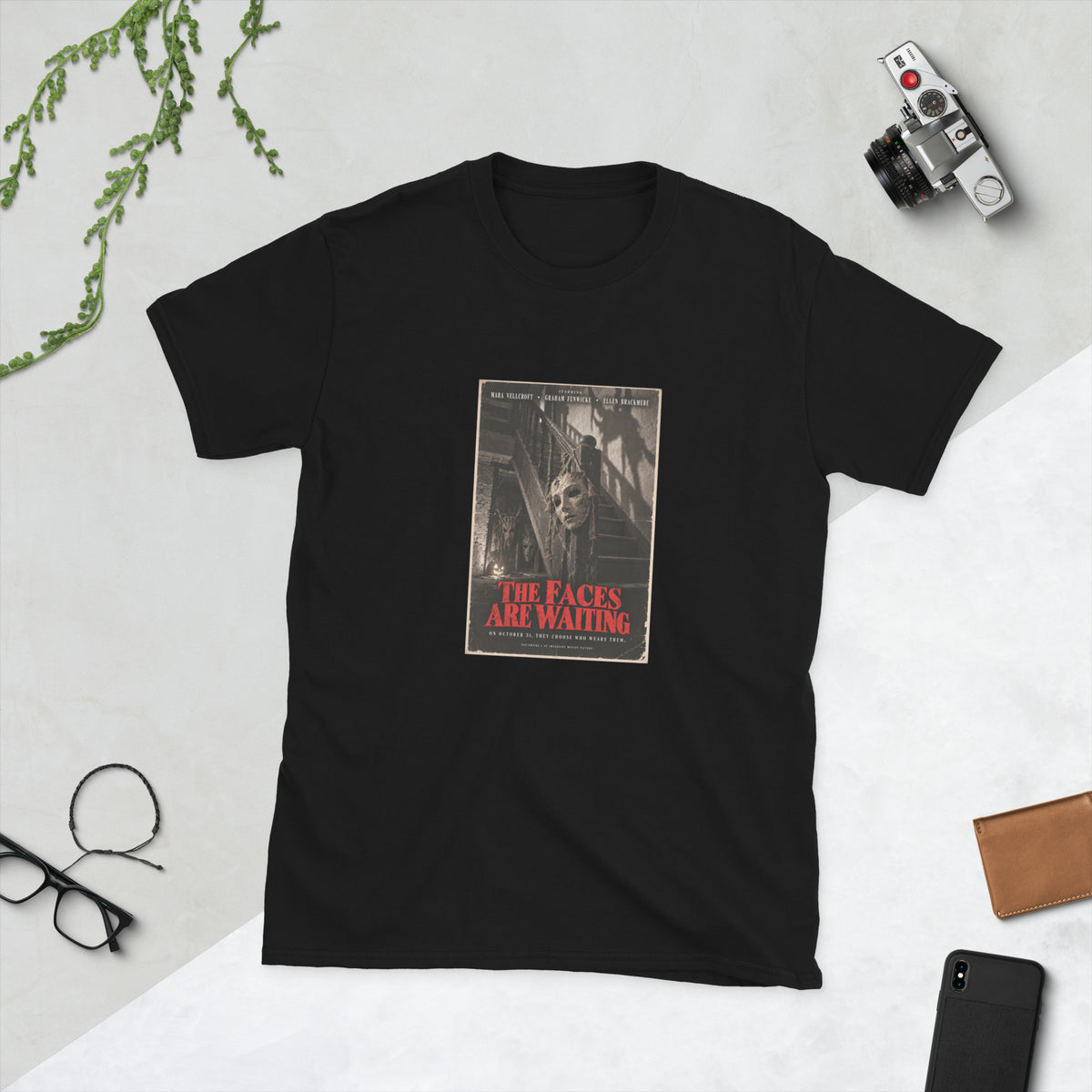 Black t-shirt printed with a sepia horror movie poster of a cracked plaster mask hanging by twine from a staircase post, with two more masks and a jack-o'-lantern in the shadows below, under the red title "The Faces Are Waiting."