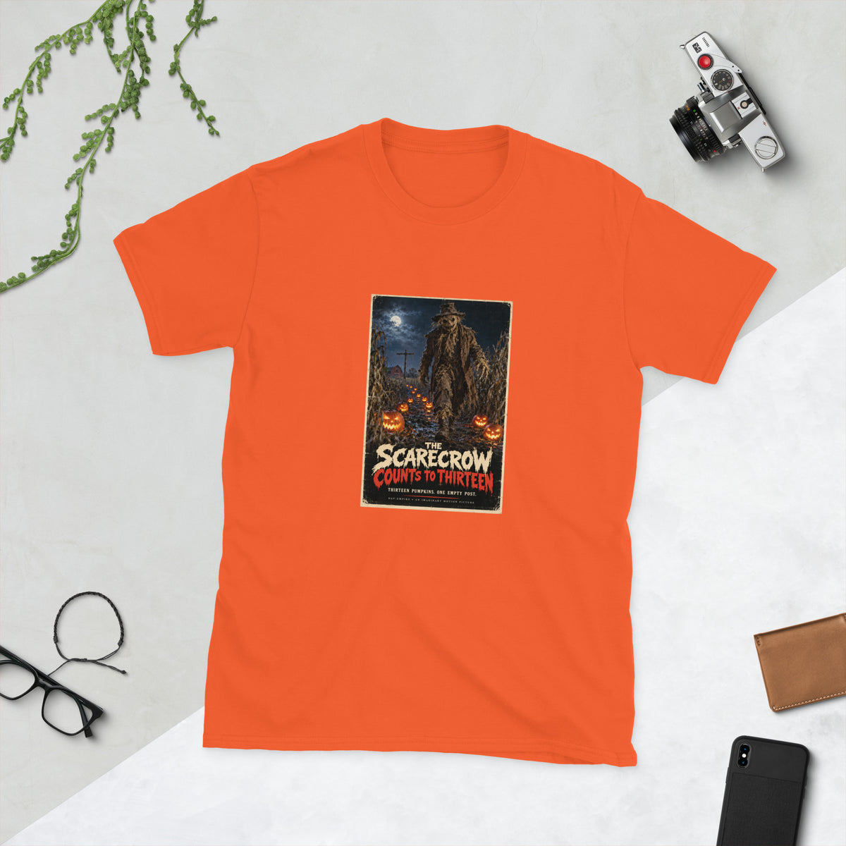 Orange t-shirt printed with a horror movie poster of a tattered scarecrow walking down a muddy cornfield path lined with glowing jack-o'-lanterns, with a red barn and an empty wooden post under a full moon behind it, titled "The Scarecrow Counts to Thirteen."
